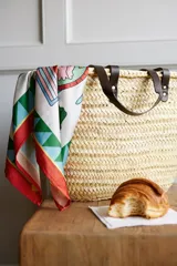 Bolso tipo capazo de rafia color natural, con asas de cuero negro y pañuelo estampado atado en una de las asas.