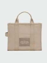 Bolso tote mediano color beige con estampado frontal del logo "The Tote Bag Marc Jacobs" en blanco. Tiene doble asa corta y correa larga extraíble de tela azul marino.
