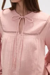 Blusa holgada de algodón color rosa pálido, con escote redondo y lazo ajustable en el centro. Presenta detalles de encaje calado alrededor del escote y en los puños de las mangas largas abullonadas.