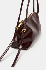 Bolso de hombro tipo baguette, color borgoña oscuro con acabado charol brillante. Presenta un asa delgada ajustable con hebilla metálica dorada y cierre superior de cremallera.