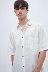 Camisa de manga corta en tela de algodón color blanco con rayas verticales en relieve y bolsillo en el pecho.