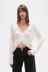 Sweater de poliéster color beige, con cuello en V, manga larga y frunces ajustables en el frente. Presenta un diseño de tejido calado.