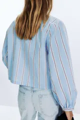 Camisa celeste con rayas verticales marrones y blancas, cuello redondo con escote en pico y mangas largas abullonadas. Cierre frontal con lazos con pespuntes marcados.