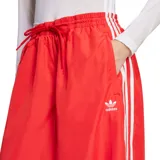 Pantalón Adidas rojo de corte ancho con cintura elastizada y cordón ajustable. Presenta las tres tiras blancas características de la marca en los laterales y el logo bordado en la pierna izquierda.