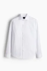 Camisa blanca de algodón con cuello italiano, tapeta lisa, canesú en la espalda y bajo redondeado. Mangas largas con tapeta de manga y botón, y cierre ajustable de botón en los puños. Corte ajustado que realza el contorno del cuerpo creando una silueta entallada.