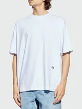 Remera blanca de manga corta y cuello redondo, con corte holgado y un pequeño parche rectangular negro con el logo de Diesel en la parte inferior delantera.