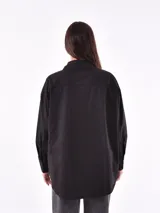 Camisa negra de corte recto, con cuello clásico, cierre frontal con botones y mangas largas con puños abotonados. Presenta dos bolsillos de parche en el pecho con apliques de tachas blancas formando un patrón geométrico.