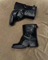 Botas negras de caña media (20 cm) de cuero vegano, con suela track gruesa y detalle de hebilla ajustable en la parte superior. Presentan un diseño arrugado o 'slouchy' en la caña.