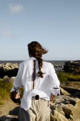 Camisa blanca de corte masculino con detalles femeninos. Presenta puños con botones dorados y cinta de terciopelo negra, y un juego de cintas verticales en contratono en la espalda. El ruedo trasero es redondeado con tablón.