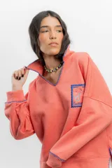 Buzo color coral de algodón con cuello polo y bordados azules en cuello, puños y ruedo. Presenta un bordado en el frente con un diseño de sirena y texto.