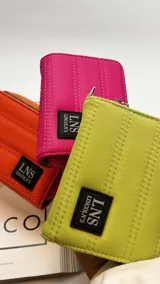 Billetera corta con cierre alrededor y tapa, confeccionada en tela capitoneada. Incluye varias reparticiones con tarjeteros y bolsillos internos, y una correa de mano desmontable. Disponible en negro, rojo y dorado.