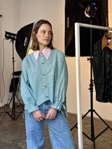 Camisa de popelina a rayas verticales verdes y blancas, con cuello rosa y botones marmolados. Es más corta adelante y más larga atrás.