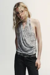 Top halter de tul color gris claro, con escote fluido drapeado y espalda descubierta. Presenta bordados con aplicación de abalorios en tonos oscuros y dorados.