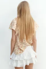 Blusa de tejido crepé color crema con estampado de flores pequeñas en tonos rosados y marrones. Tiene corte holgado, cuello con volados y escote en V con tiras para ajustar. Las mangas son cortas tipo mariposa con ribete de volado.