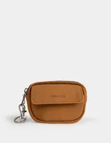 Monedero color marrón camel de suede con bolsillo frontal con solapa y broche con botón para cerrado. Tiene cierre superior con mosquetón y tag en relieve con la inscripción "Girls Club".