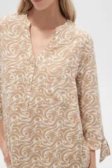 Blusa color beige con estampado de arabescos blancos, cuello mao, mangas 3/4 con lazos en los puños, cierre parcial con botones y bolsillos frontales.