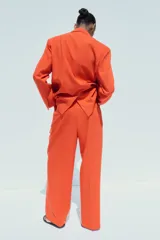 Pantalón de vestir color naranja, de tiro alto y pernera ancha. Presenta doble pliegue frontal, trabillas en la cintura, bolsillos laterales y falsos bolsillos de vivo en la espalda.