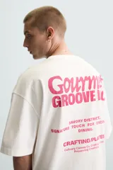Camiseta blanca de corte holgado, cuello redondo y manga corta. Estampado frontal con la frase "Gourmet Groove LLC" en color fucsia.