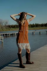 Vestido corto marrón de gamuza sintética, sin mangas, con escote en V y detalle de flecos en el ruedo. Presenta apliques bordados en el frente y laterales.