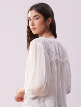 Blusa color beige de gasa con cuello a la base, escote en V y abertura central con botones. Presenta detalle de crochet en hombros y espalda. Manga 3/4 amplia con puño y botón.