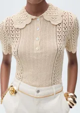 Jersey de punto calado color beige, con cuello polo, terminación en ondas, manga corta y cierre de botones en el cuello.