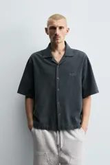 Camisa de denim de algodón, color beige claro, con corte relajado, cuello bowling y manga corta. Presenta cierre frontal con botones a presión y un pequeño bordado a tono en el pecho.