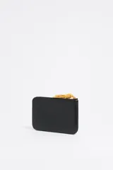 Monedero negro de cuero con cierre de cremallera y tirador de cordón amarillo. Incluye bolsillo interior y placa metálica con el logo de la marca.