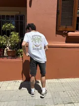 Remera blanca de manga corta con estampa en la espalda de dos palmeras, una persona en una hamaca y el texto 'Paradise Holiday Brixton'.