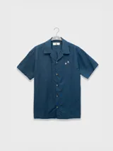 Camisa azul de hombre de calce relajado y corte cuadrado, confeccionada en una mezcla liviana de lino y algodón. Presenta cuello tipo camp, cierre con botones de madera grabados con el logo y un bordado sutil en el pecho izquierdo con forma de palmera. Incluye patch con logo de la marca.
