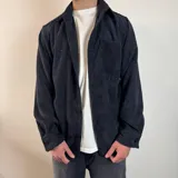 Chaqueta tipo sobrecamisa de pana color negro, con cuello clásico, cierre frontal abotonado, bolsillo de parche en el pecho y corte recto.