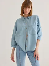 Camisa de denim azul con pespuntes blancos, corte crop y cierre frontal con botones. Tiene cuello clásico, mangas cortas y un bolsillo de parche en el pecho. Se ata en la parte delantera.