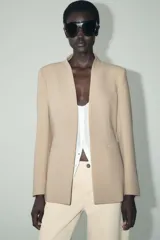 Blazer color beige, sin solapa, de manga larga con bolsillos de vivo en el delantero y cierre frontal con gancho metálico.