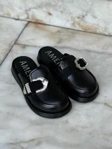 Zuecos negros de cuero con suela gruesa, detalle de tira de gamuza con hebilla metálica plateada y logo "AMÉN" estampado en la plantilla.