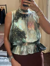Blusa sin mangas con cuello alto fruncido y peplum elástico en la cintura. Presenta un estampado abstracto en tonos oscuros (negro, verde azulado, rosado) con detalles de puntos dorados metalizados.