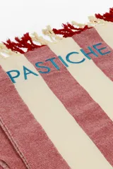 Pareo-lona rectangular con rayas horizontales rojas y blancas, con flecos en los extremos y la palabra "PASTICHE" bordada en azul.