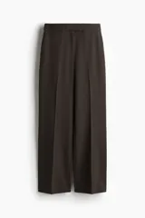 Pantalón de sarga color marrón oscuro, de tiro alto con elástico oculto en la cintura, cierre y pretina prolongada con botón y corchetes ocultos. Tiene piernas con corte para bota, bolsillos al bies, bolsillos de atrás ribeteados y pliegues adelante y atrás.