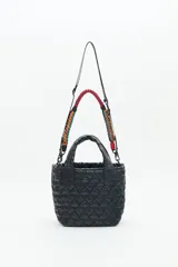 Cartera tote azul de nylon matelassé con diseño de corazones. Tiene manijas cortas y dos correas desmontables, una de cuero sintético y otra de tela trenzada multicolor. Cuenta con cierre superior y dos bolsillos internos.