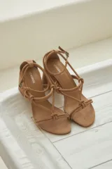 Sandalias color tostado confeccionadas en cuero, con tiras finas y detalle de nudos en el empeine. Ajuste de pulsera en el tobillo y taco recto de 7cm.