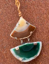Collar con colgante de dos piedras naturales irregulares engarzadas en metal plateado y dorado, suspendidas de un cordón de cuero marrón con detalle de nudo corredizo. Las piedras tienen vetas en tonos naranja, marrón y blanco.