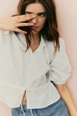 Blusa blanca de algodón con textura de lunares. Tiene escote redondo con lazo, botones en el frente, mangas tres cuartos abullonadas, cordón ajustable en la cintura y peplum.