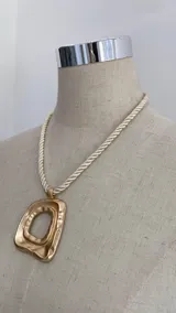 Collar con cadena de cordón trenzado color crema y dije dorado con diseño abstracto irregular.