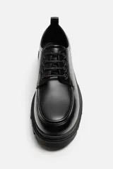 Zapato de vestir tipo blucher color negro, con detalle de bordón, tirador trasero y horma redonda. Suela con volumen y diseño irregular.