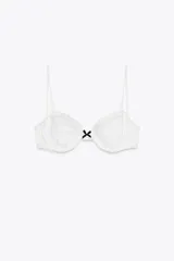 Top de bikini blanco con aros y tirantes finos ajustables. Tiene volantes en el pecho y un lazo negro en el centro. Cierre trasero con gancho metálico.