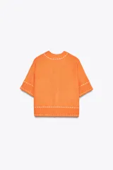 Chaqueta naranja de punto con cuello redondo y manga corta. Presenta bolsillos delanteros de parche con solapa y cierre frontal con botones. Detalle de pespuntes blancos a contraste.