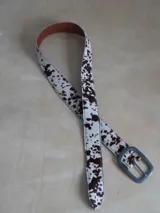 Cinturón de cuero sintético con estampado animal print de vaca en tonos marrones y blancos. Hebilla metálica plateada con logo grabado.