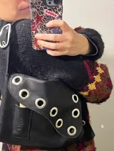 Cartera bandolera de cuero negro con ojalillos metálicos plateados en el frente y correa regulable con hebilla. Tiene un bolsillo exterior y gancho interior para llaves.