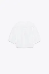 Camisa blanca de popelín con cuello bobo bordado y lazada. Tiene mangas abullonadas por debajo del codo con elástico y pinzas en la espalda. El cierre frontal es con lazadas.