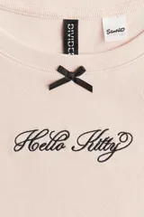 Remera rosa de algodón con cuello redondo y mangas cortas. Presenta un pequeño moño negro y el logo de Hello Kitty bordado en el pecho.