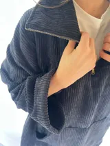 Buzo de pana fina color verde oliva, con diseño de corte holgado y mangas largas. Presenta una textura de cordón vertical característica de la pana y un pequeño parche decorativo en la parte superior de la espalda.