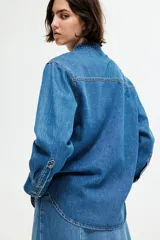 Camisa de denim azul de corte holgado, con cuello mao, tapeta de botones, hombros caídos, mangas largas con puños abotonados y dos bolsillos superiores con solapa.